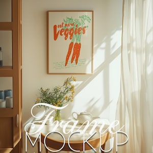 Puede incluir: Un póster enmarcado con fondo blanco y texto verde que dice "eat more veggies". El póster presenta un dibujo de cuatro zanahorias naranjas con hojas verdes. El póster está colgado en una pared blanca con una mesa de madera y un jarrón de flores amarillas delante.