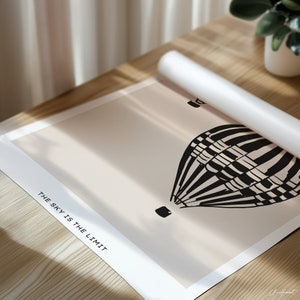 May include: A rolled-up poster featuring a black and white hot air balloon illustration against a beige background. The text "THE SKY IS THE LIMIT" is printed in black along the bottom edge. The poster is on a wooden surface.