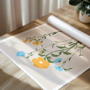 May include: A floral art print featuring a bouquet of stylized flowers in yellow and blue against a light beige background. Green stems and leaves complement the blooms. A rolled-up white poster is partially visible. The artwork is on a wooden surface.