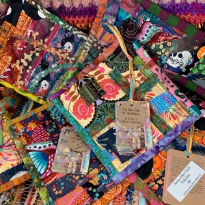 May include: A collection of colorful patchwork mini quilts with various patterns and designs. The quilts are made with different fabrics and textures, including floral prints, geometric patterns, and animal motifs. Each quilt has a tag attached with the artist's name and the title of the piece. The quilts are displayed on a woven rug with a variety of colors and textures.