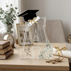 May include: Clear acrylic graduation decoration featuring a black mortarboard, diploma, and floral accents. The design includes the text "Class of 2020", "Western University", and a personalized name. A decorative graduation gift.