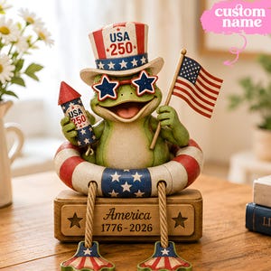 Könnte beinhalten: Eine grüne Froschfigur mit einem rot-weiß-blauen Zylinder mit der Aufschrift USA 250 und sternförmigen Brillen. Der Frosch hält ein Feuerwerk und eine US-Flagge, sitzt in einem Rettungsring auf einem Holzsockel mit der Gravur America 1776-2026.