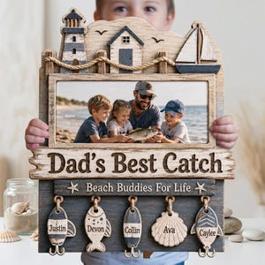 Puede incluir: Un marco de fotos de madera para pared con temática náutica que incluye un faro, un velero y un detalle de cuerda. El marco muestra una foto familiar pescando, el texto "Dad's Best Catch" y "Beach Buddies For Life", y cinco etiquetas colgantes con forma de pez con los nombres Justin, Devon, Collin, Ava y Caylee.
