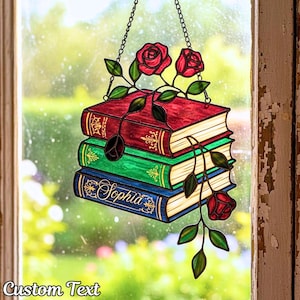 以下が含まれることがあります： 赤、緑、青の3冊の本が積み重ねられたステンドグラスの窓飾り。本はバラと葉で飾られており、一番下の本には「Sophia」という名前が書かれています。「Custom Text」という文字が下にあります。