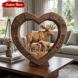 Peut inclure: Une sculpture en bois en forme de cœur représentant un orignal sculpté et deux petits cerfs. La sculpture porte les mots "DAD", "OLIVIA" et "ETHAN". Le haut du cœur est orné de lanières de cuir et de rivets en laiton. Le texte "Custom Name" est en haut.