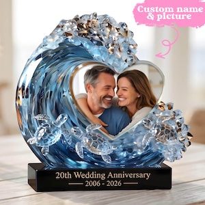 Può includere: Una cornice per foto a forma di onda blu e trasparente con apertura a forma di cuore. La cornice presenta la foto di una coppia, tartarughe marine e il testo "20° anniversario di matrimonio 2006 - 2026". In alto c'è il testo "Nome e immagine personalizzati".