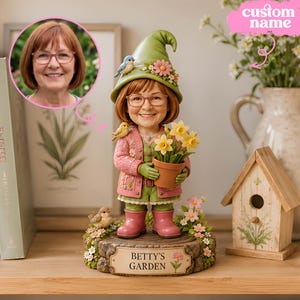 Könnte beinhalten: Eine personalisierte Figur einer Person mit braunen Haaren und Brille, die einen grünen Hut, eine rosa Strickjacke und rosa Stiefel trägt. Die Figur hält einen Terrakottatopf mit gelben Blumen und steht auf einem Sockel mit der Aufschrift BETTY'S GARDEN.