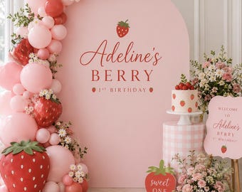 以下が含まれることがあります： Adeline's BERRY 1ST BIRTHDAYという文字とイチゴのグラフィックが描かれたピンク色のアーチ型背景。ピンクと赤のバルーンガーランド、イチゴ型の装飾、ケーキ、sweet ONEと書かれた看板で飾られています。