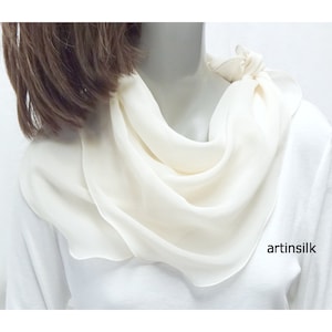 May include: A cream-colored silk scarf draped around a mannequin's neck. The scarf is soft and flowing, with a subtle sheen. The fabric is gathered and knotted at the back, creating a layered effect. The scarf is a square shape.
