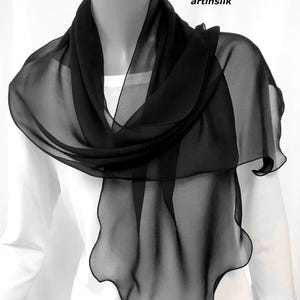 May include: A sheer black scarf draped over a white top. The scarf is made of a lightweight, flowing fabric, with a wavy, scalloped edge. The text "artinsilk" is visible in the upper right corner.