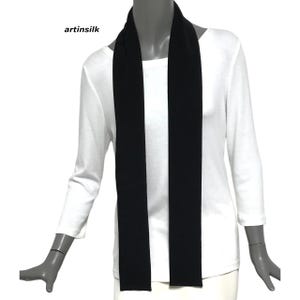 May include: A black scarf draped over a white long-sleeved shirt. The scarf is a solid black color and hangs vertically. The text "artinsilk" is visible in the upper left corner. The scarf appears to be made of a soft material.