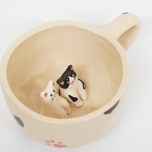 Puede incluir: Taza de cerámica color crema con asa, con dos pequeñas figuras de gato en su interior. Un gato es blanco con marcas negras, el otro es blanco. La taza tiene lunares negros y huellas de patas.