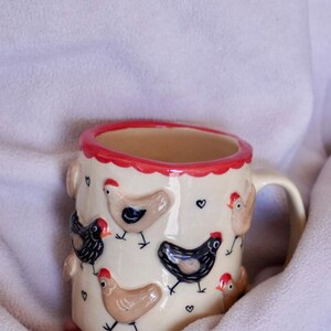 Op de afbeelding: Crèmekleurige keramische mok versierd met reliëfkippen in zwart, bruin en bruin, elk met een rode kam. De rand is rood en kleine zwarte hartjes zijn verspreid rond de mok. Een charmant, grillig ontwerp voor een keuken of boerderijdecor.