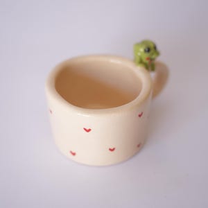 Op de afbeelding: Een kleine, cr&egrave;mekleurige keramische mok met rode hartvormige ontwerpen. De mok heeft een klein handvat en een groene kikkerfiguur op de rand. De mok wordt waarschijnlijk gebruikt voor het drinken van dranken.