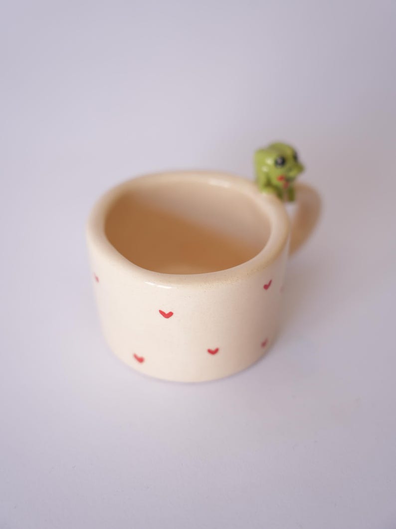Op de afbeelding: Een kleine, cr&egrave;mekleurige keramische mok met rode hartvormige ontwerpen. De mok heeft een klein handvat en een groene kikkerfiguur op de rand. De mok wordt waarschijnlijk gebruikt voor het drinken van dranken.