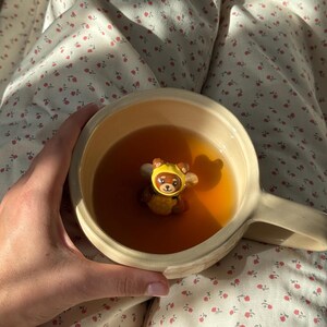 Puede incluir: Una taza de cerámica de color crema llena de té de color ámbar, con una pequeña figura tridimensional de un oso marrón que lleva una capucha y alas con temática de abeja, situada en el fondo de la taza y parcialmente sumergida.