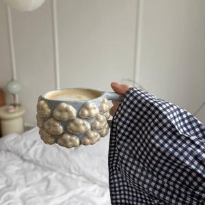 Könnte beinhalten: Eine hellblaue Keramiktasse mit Wolkenmuster. Die Tasse ist mit einem cremigen Getränk gefüllt. Die Tasse wird von einer Person gehalten, die ein schwarz-weiß kariertes Hemd trägt. Die Tasse hat einen Henkel.
