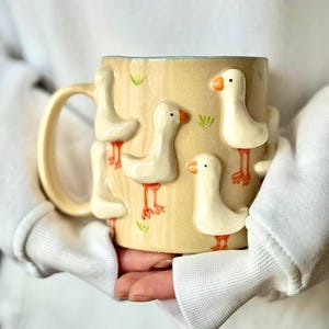 May include: A beige ceramic mug featuring raised relief goose figures with orange beaks and feet. The mug has a light blue interior and a matching handle. Green grass accents are painted around the geese.