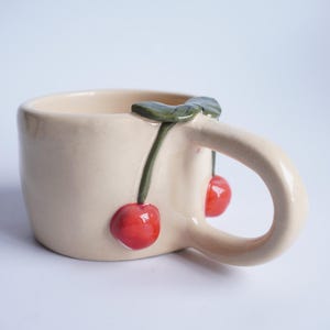 Puede incluir: Taza de cerámica color crema con un asa redondeada, adornada con dos cerezas rojas y hojas verdes. Las cerezas están unidas a la taza con tallos verdes. La taza tiene un acabado liso y brillante sobre un fondo blanco.