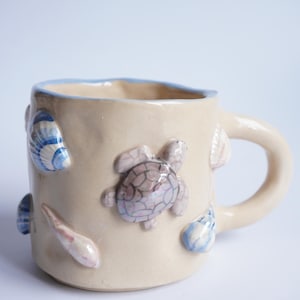 May include: Cream ceramic mug with a light blue rim and handle. The mug is decorated with raised seashell and turtle designs in blue, pink, and white. A unique, handmade piece for enjoying hot drinks, ideal for the home.