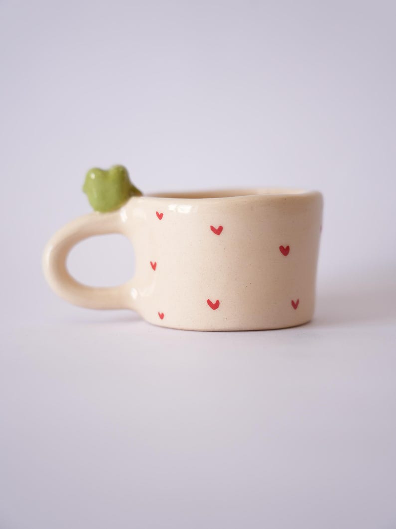 Op de afbeelding: Cr&egrave;mekleurige keramische mok met rode hartaccenten. Een kleine groene kikker siert het handvat. De mok is ontworpen voor dranken en heeft een afgerond handvat voor gemakkelijk vasthouden. Een charmante toevoeging aan elke keuken.