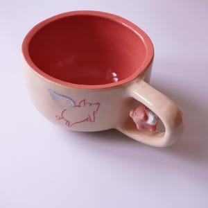 Könnte beinhalten: Eine Keramik-Tasse mit pfirsichfarbenem Innenraum und beigem Äußeren. Die Tasse zeigt eine rosa fliegende Schweine-Illustration und eine kleine Schweinefigur am Griff. Die Tasse ist für Getränke konzipiert.