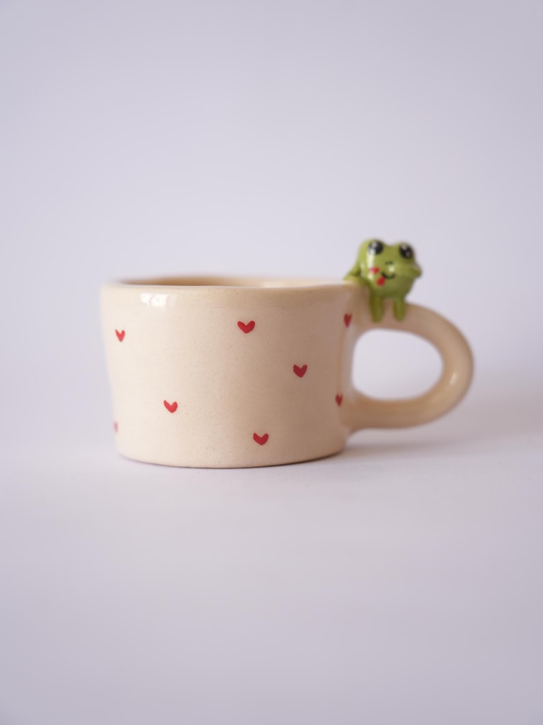 Op de afbeelding: Een cr&egrave;mekleurige keramische mok versierd met kleine rode hartjes. Een groene kikkerfiguur zit op de rand. De mok heeft een afgeronde handgreep en een licht uitlopende vorm, ideaal voor koffie of thee.