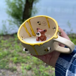 Op de afbeelding: Een grillige keramische mok met een berenfiguur erin, omringd door een bruisende vloeistof. De mok heeft een crèmekleurige basis met gele honingachtige druppels en bijenaccenten. De handgreep is ook crèmekleurig. De achtergrond toont een wazige buitenscène.
