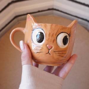 Puede incluir: Una taza de cerámica con forma de cabeza de gato atigrado naranja con ojos grandes y separados, pestañas largas, una pequeña nariz rosa y bigotes pintados. La taza presenta dos orejas puntiagudas en el borde superior y un asa curva.