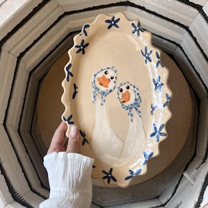 May include: An oval ceramic platter with a scalloped edge, featuring a cream-coloured glaze and a hand-painted pattern of blue flowers along the rim. The centre displays two white geese wearing blue and white patterned headscarves.