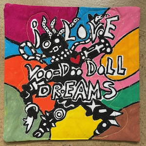 May include: A square fabric pillow cover featuring a colorful, abstract background of pink, blue, orange, yellow, and green shapes. A black, stylized figure is centered with the text "I LOVE VOODOO DOLL DREAMS" and a small red heart.