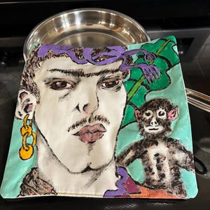 May include: Square, hand-painted pot holder featuring a stylized portrait of a person with a mustache, gold earring, and a monkey. The background is light blue with green leaves and purple accents. The pot holder is made of fabric.