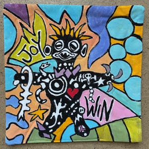 May include: A square fabric cushion cover featuring a vibrant, hand-painted illustration of a stylized black figure with yellow hair. The design includes the words JOY and WIN, a red heart, stars, and abstract shapes in blue, orange, and yellow tones.