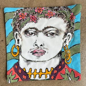 May include: Square fabric art featuring a portrait of a person with a floral crown, gold hoop earrings, and a yellow necklace. The background has blue and green stripes. The person's shirt is red with purple and orange accents.
