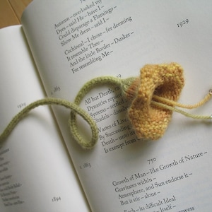May include: A yellow knitted flower bookmark with a green cord. The bookmark is on an open book with text about death and nature. The text on the book includes the years 1863, 1894, 1929, and 750.