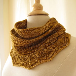 May include: Mustard-colored knitted cowl with intricate stitch patterns. The cowl is displayed on a mannequin, showcasing its textured design and triangular shape. The warm, earthy tone of the cowl suggests a cozy accessory.