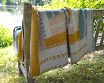 May include: A knitted throw blanket with a geometric pattern in shades of gray, yellow, and blue. The blanket is draped over a wooden bench.