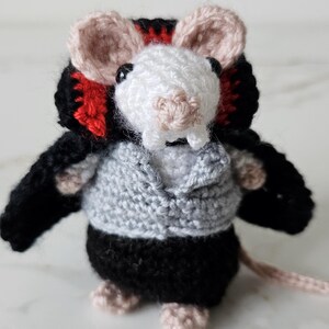 Könnte beinhalten: Eine handgefertigte Häkelmauspuppe mit weißem Kopf, rosa Ohren und einem grauen Pullover. Die Maus trägt eine schwarz-rote Kopfbedeckung und einen schwarzen Unterteil. Die Puppe hat einen rosa Schwanz und Füße.