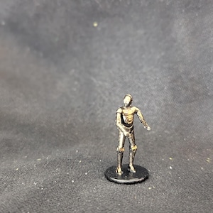 May include: A small, metallic figurine of a humanoid robot with a detailed design. The figure is silver and gold, standing on a small, black circular base. The robot has a slender build and is posed with one arm extended.