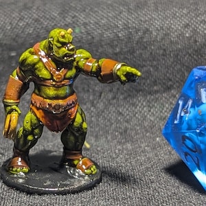 May include: A green and brown painted miniature figurine of a goblin-like creature with a horned head, wearing a red loincloth and a brown leather strap on its left arm. The figurine is standing on a black base and is pointing with its right hand.