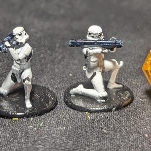 May include: Two miniature Stormtrooper figurines, one standing and one kneeling, both holding blasters. The figures are painted white with black accents and are mounted on black bases. A yellow, faceted die is in the background.