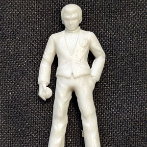 May include: A white plastic figurine of a person wearing a suit. The figurine is standing with its arms at its sides and has a neutral expression on its face.