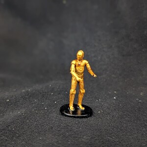 May include: A gold-colored figurine of a humanoid robot with articulated limbs, standing on a black circular base. The robot has a detailed design with visible mechanical elements. The background is a dark, textured fabric.
