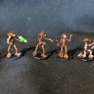 May include: A collection of miniature copper-colored robot figurines. The robots vary in design, some standing, some kneeling, and one holding a green and yellow weapon. Each figure is mounted on a small black base, creating a sci-fi or fantasy theme.