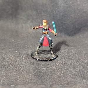 May include: Miniature figurine of a character in a dark blue and red outfit, holding a blue lightsaber. The figure is on a black base, with details in gold and brown. The character is in a dynamic pose.