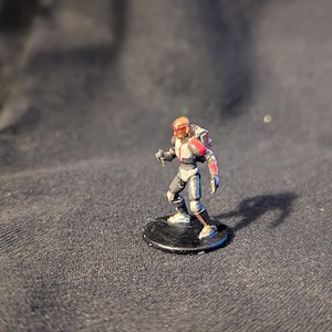 May include: Miniature figurine of a futuristic soldier in a gray and red armored suit, standing on a black circular base. The figure has a helmet and is holding a weapon. The miniature is likely for tabletop gaming.