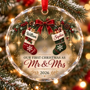 Könnte beinhalten: Ein runder, transparenter Weihnachtsbaumschmuck aus Glas mit roter Schleife, Tannenzweigen und zwei Strümpfen mit den Namen William und Sophia. Der Text lautet Our First Christmas as Mr & Mrs 2026, verziert mit goldenen Schneeflocken.