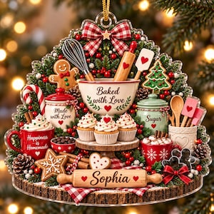 May include: A decorative Christmas ornament featuring a baking theme with a mixing bowl labeled Baked with Love, a rolling pin labeled Sophia, a gingerbread man, cupcakes, red mugs with text, a sugar jar, and festive greenery with a plaid bow.