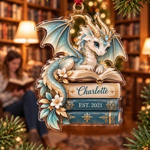 May include: A clear acrylic Christmas ornament featuring a white and blue dragon perched on a stack of books. The top book is open, while the spine of the middle book reads Charlotte and the bottom book reads EST. 2021. White flowers accent the base.