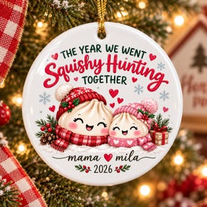 Könnte beinhalten: Ein runder, weißer Keramik-Weihnachtsbaumschmuck mit zwei lächelnden Cartoon-Teigtaschen, die rote und rosa Wintermützen und Schals tragen. Der Text lautet THE YEAR WE WENT Squishy Hunting TOGETHER, mama Herz mila und 2026.